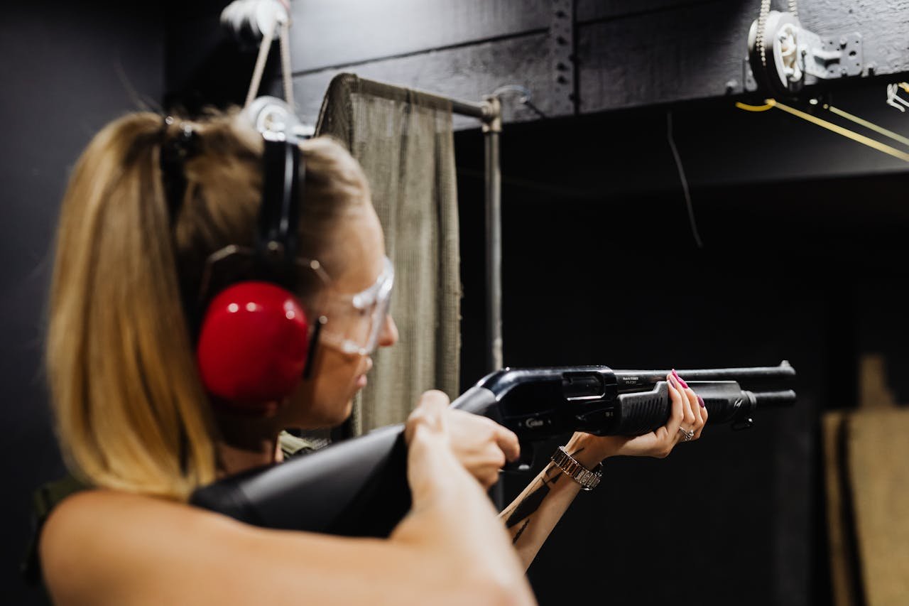 A focused young woman aims a shotgun at an indoor shooting range, wearing safety gear.